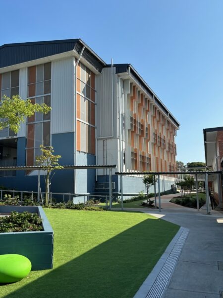 Three-storey learning building at Balmoral State School, orange accent panels and vertical sun-shade fins beside a landscaped courtyard