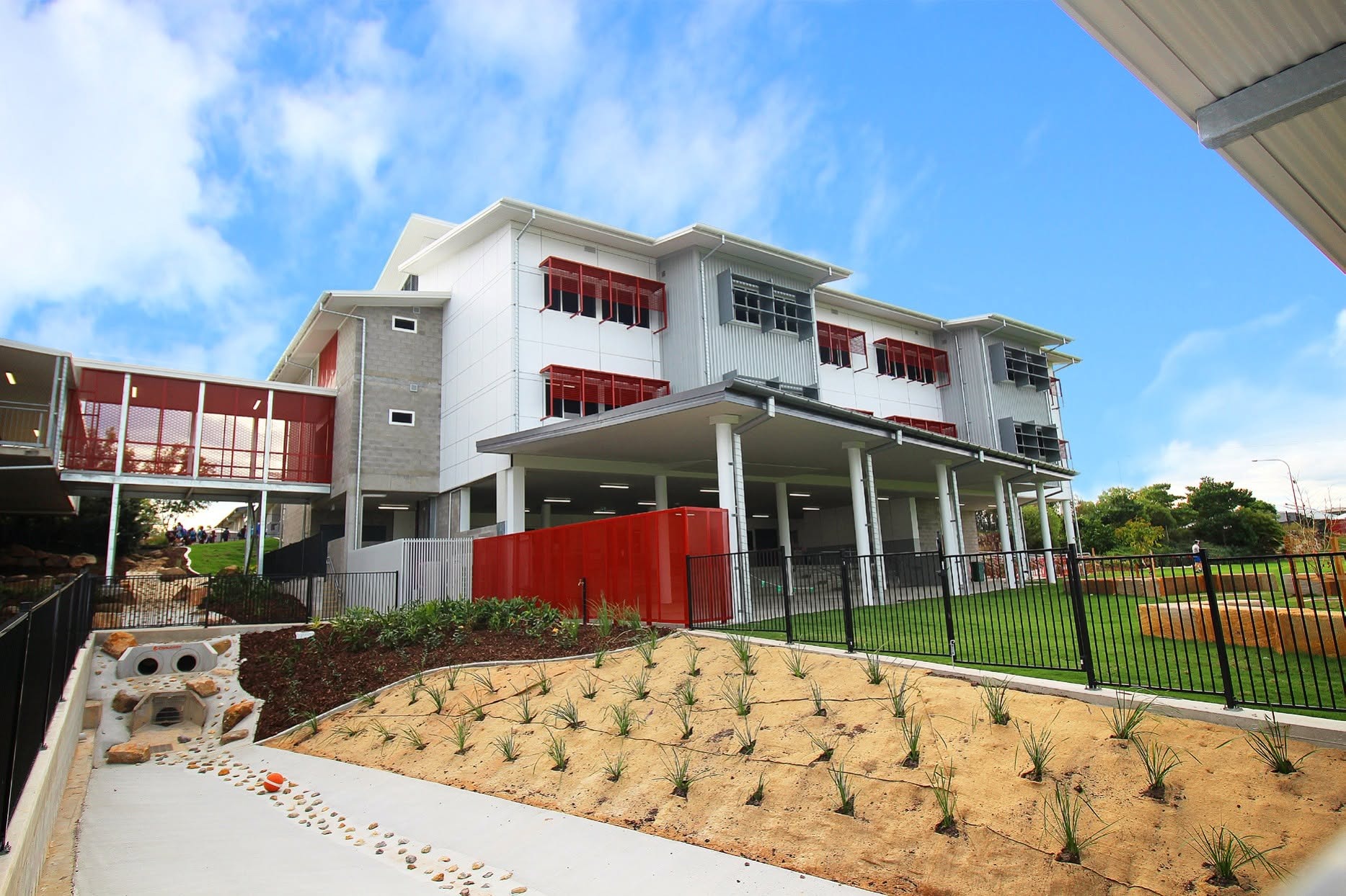 Three-storey learning building at Griffin State School, white cladding with red louvres and an elevated walkway link, on a landscaped bank