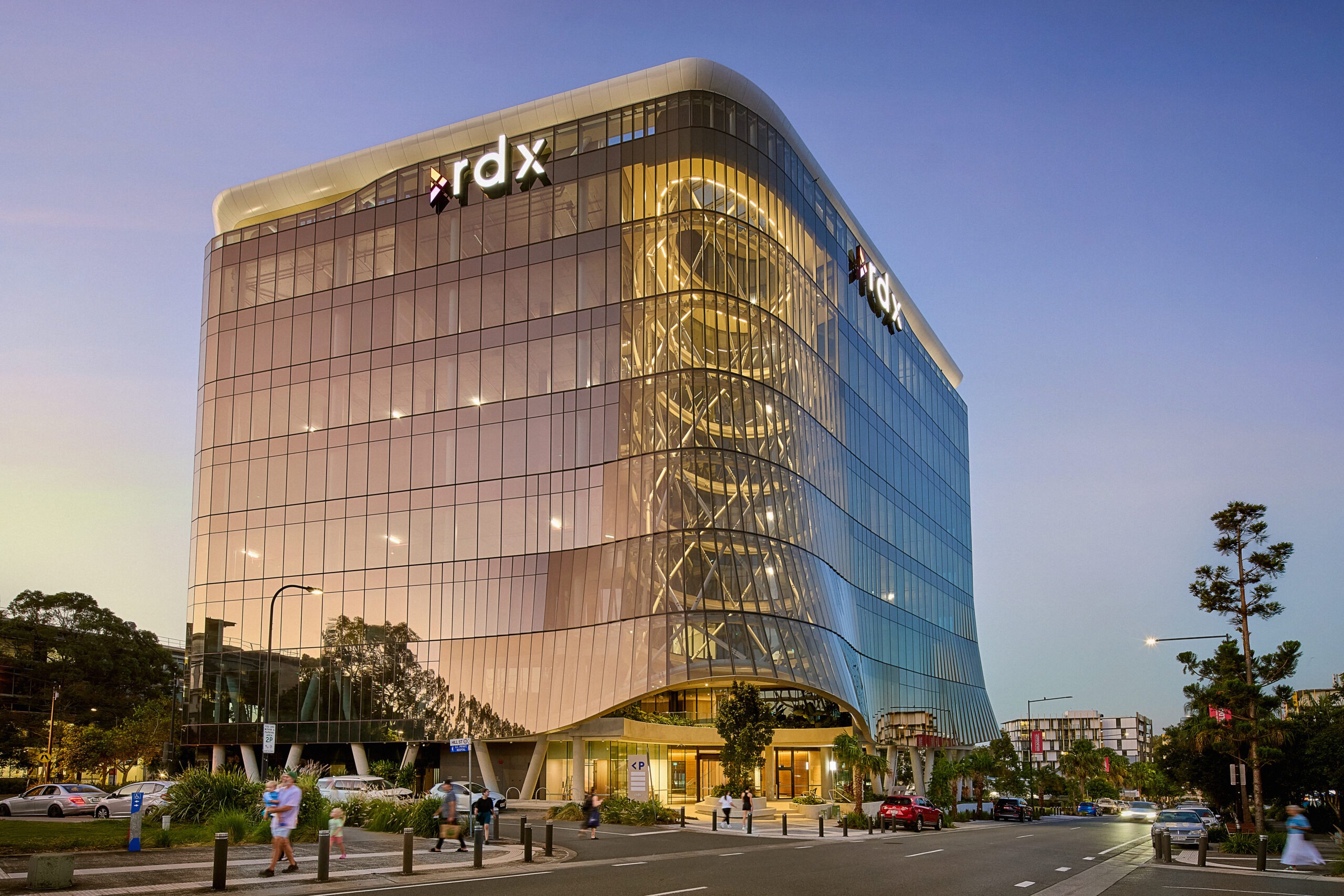 The curved bronze-glass RDX Lumina building at dusk, its full-height glazed atrium lit from within